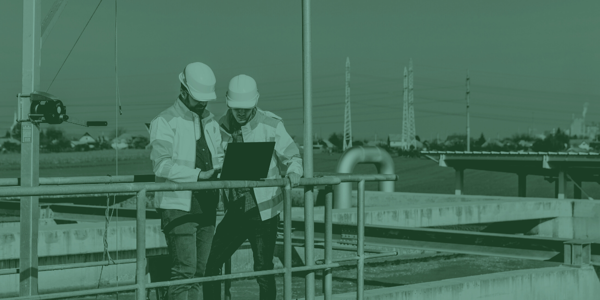 Two Cypress Fabrication workers in hard hats and safety vests at a water treatment facility looking at a laptop